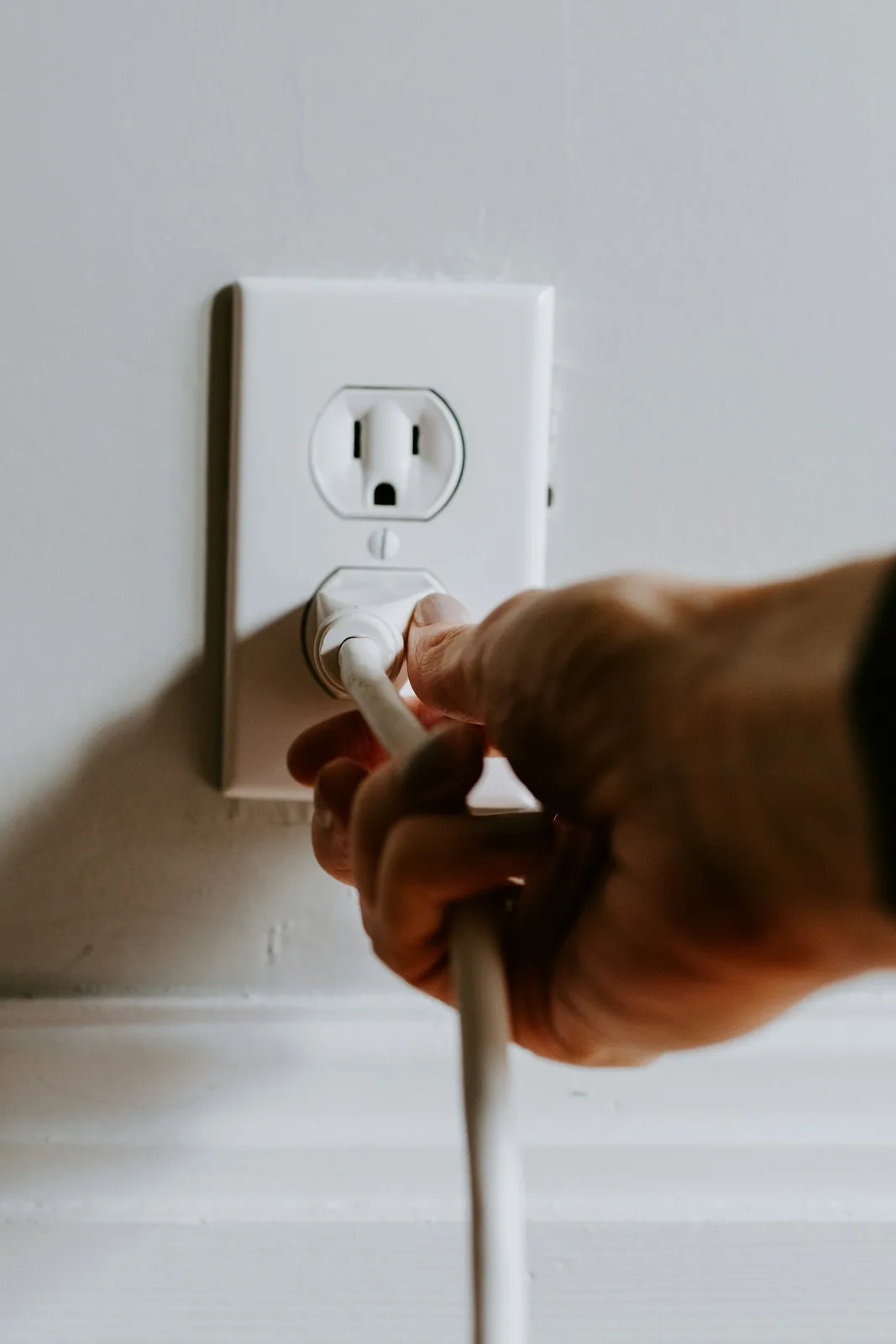 A hand inserting a white power plug into the lower socket of a US-style duplex wall outlet.