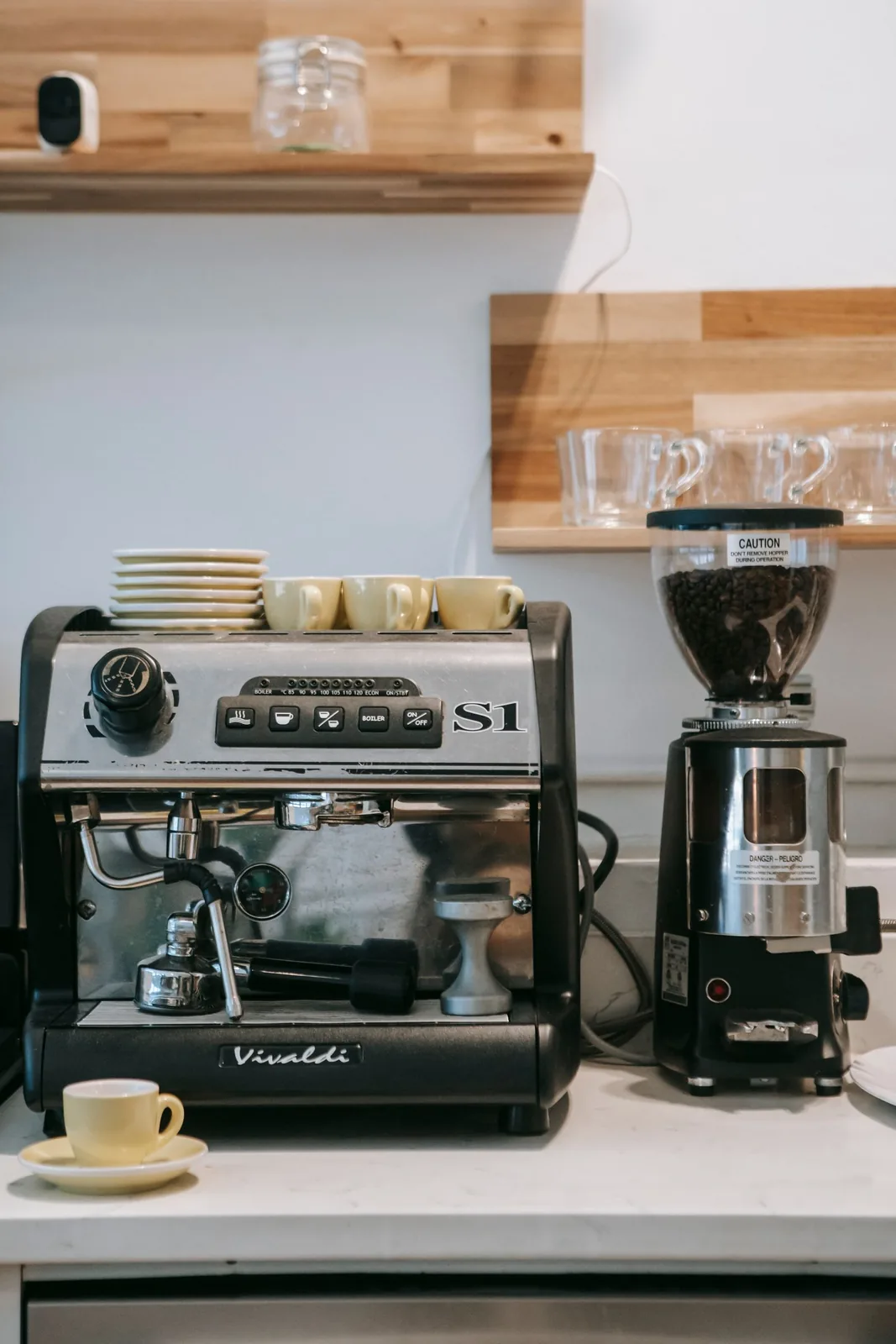 An espresso machine and separate coffee grinder on a countertop below shelves of cups.