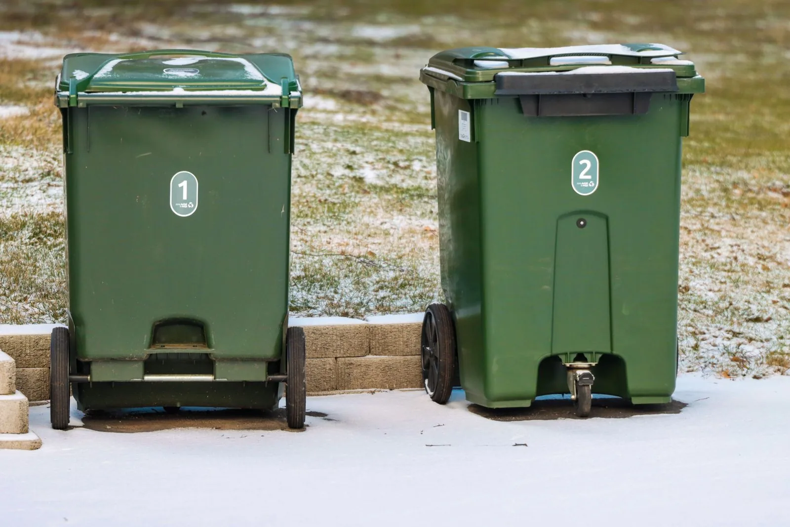 Two green wheeled waste bins standing upright on a lightly snow-covered paved area.