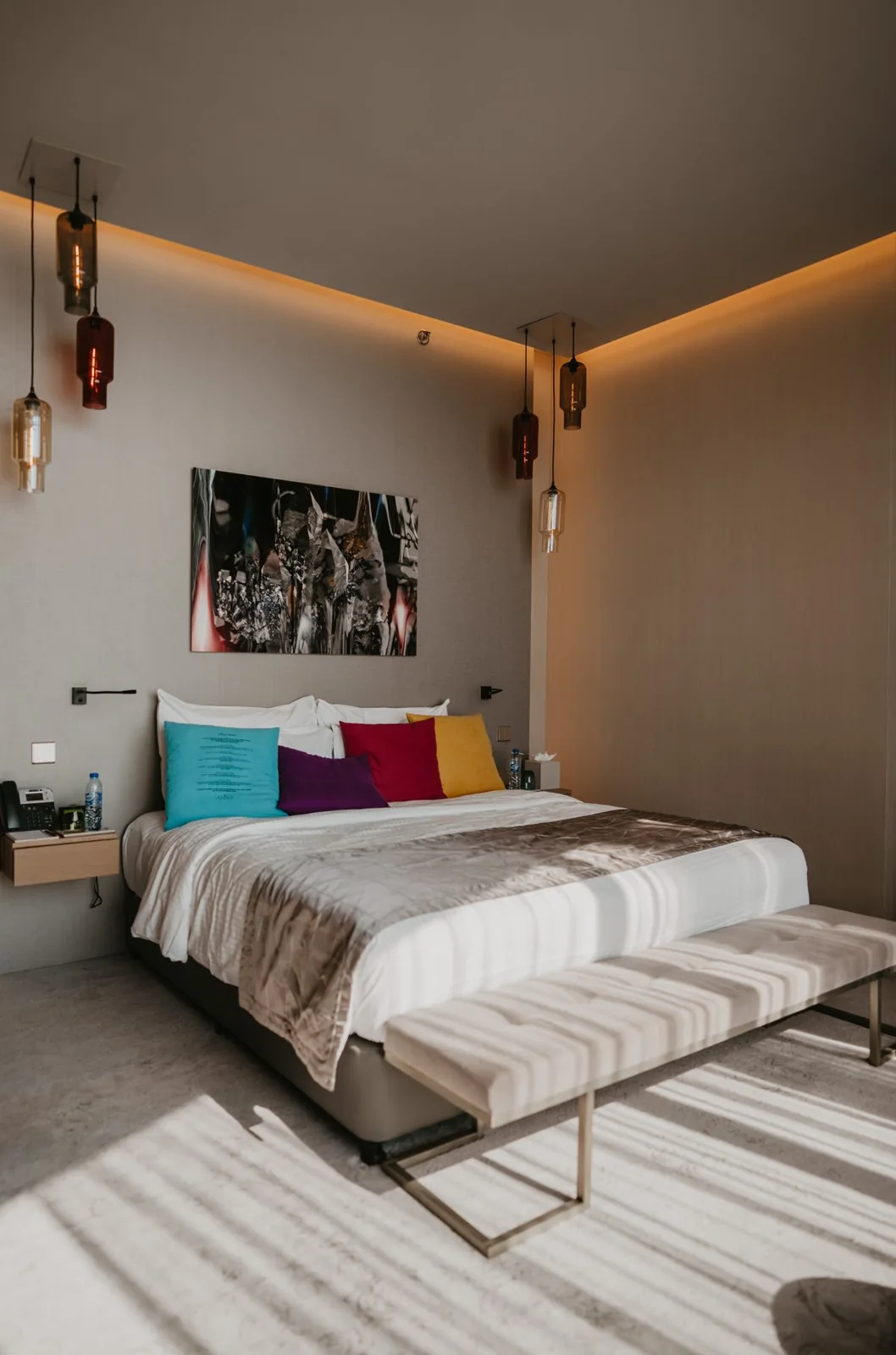 Empty guest room with a made bed, colored cushions, pendant lights and an upholstered bench.