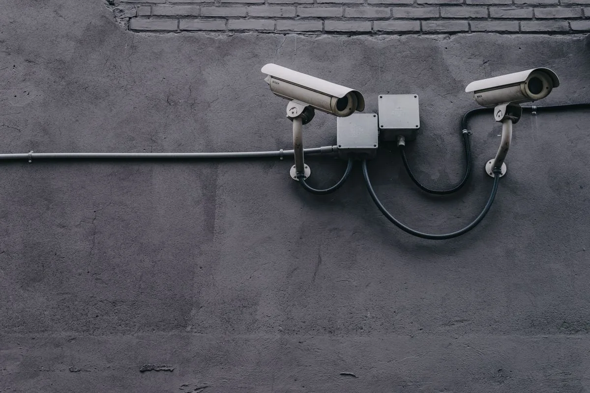 Two surveillance cameras and their cables mounted on a gray brick wall.