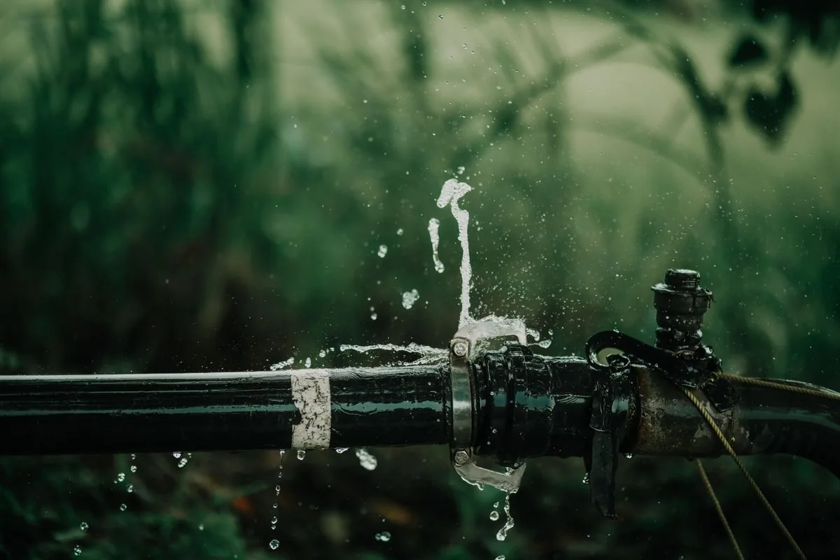 Water spraying from a joint in a black outdoor pipe against green foliage.