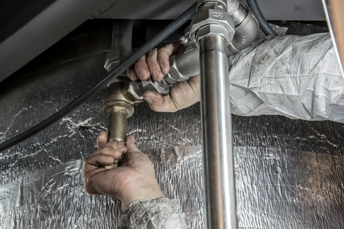 Hands working on a metal pipe connection beside insulated pipework.