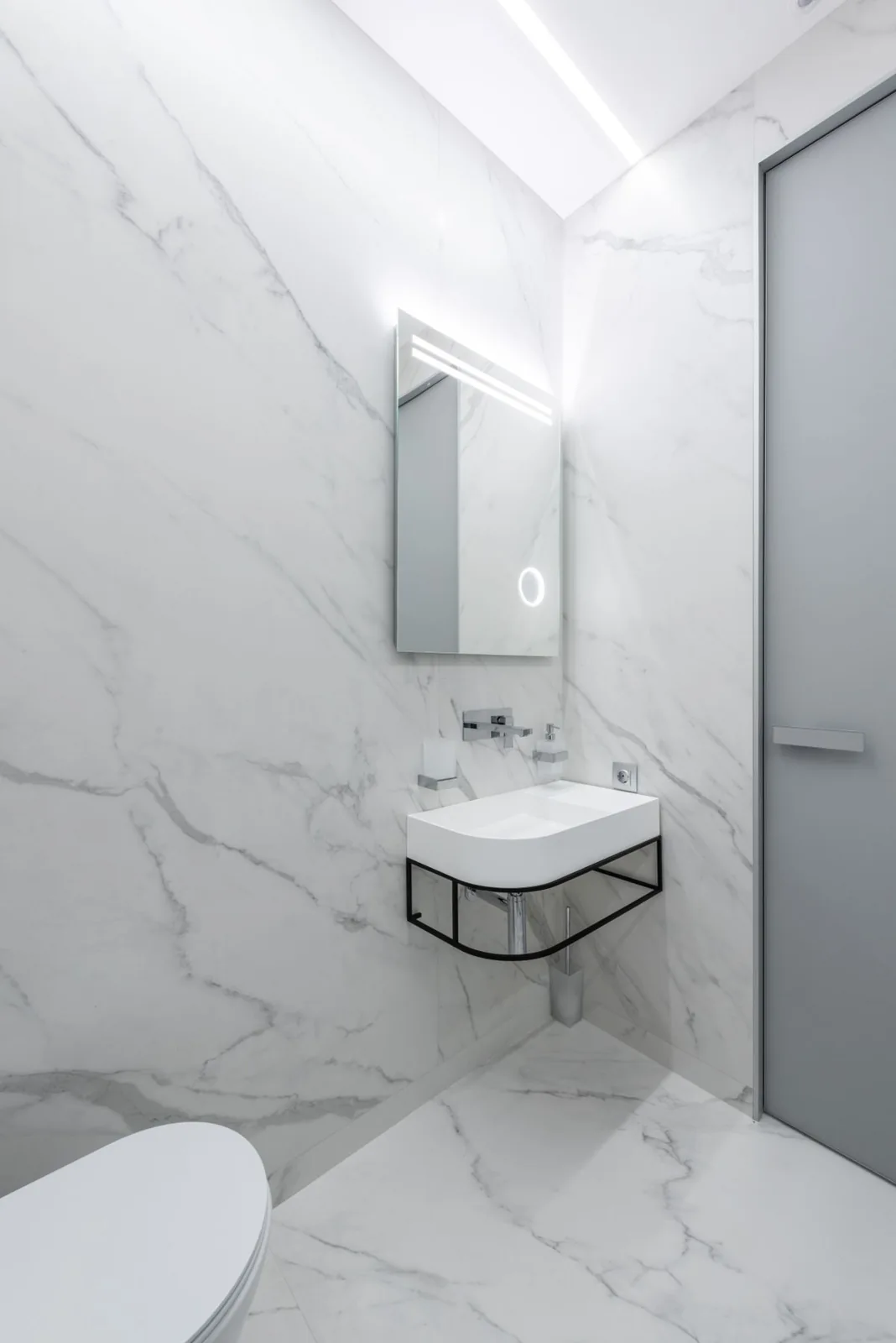 Empty white marble bathroom with a wall-mounted sink, exposed drain pipe, mirror and closed door.