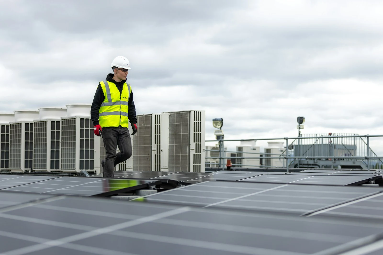 A worker in safety equipment walks between rooftop solar panels and a bank of commercial HVAC units.
